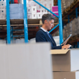Warehouse manager using a tablet to inspect inventory, representing the use of warehouse management system software in a modern distribution facility.