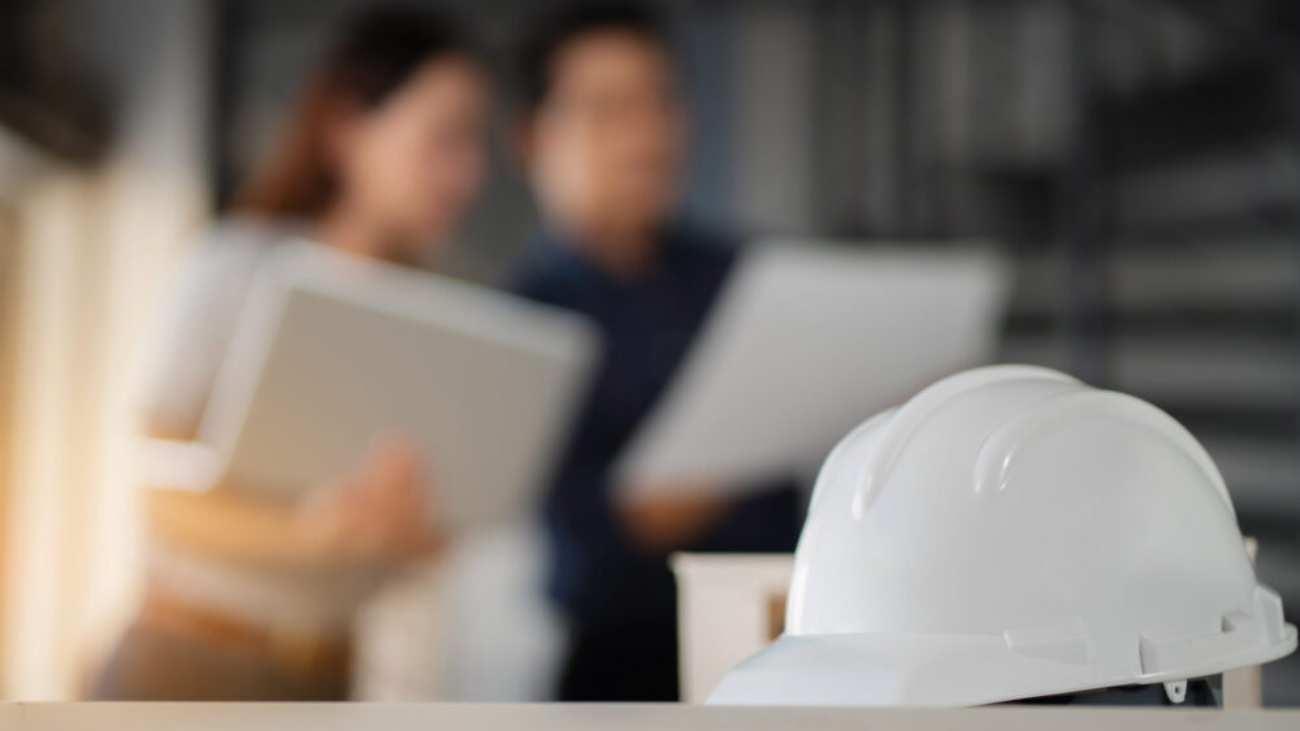 White hard hat on a desk with construction professionals discussing plans in the background, symbolising the benefits of construction ERP software.