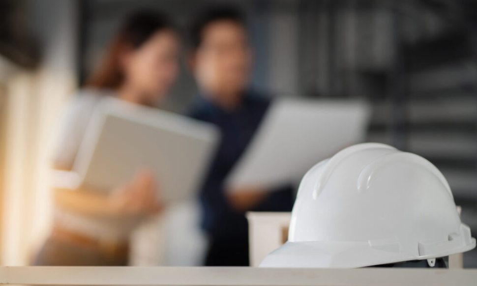 White hard hat on a desk with construction professionals discussing plans in the background, symbolising the benefits of construction ERP software.
